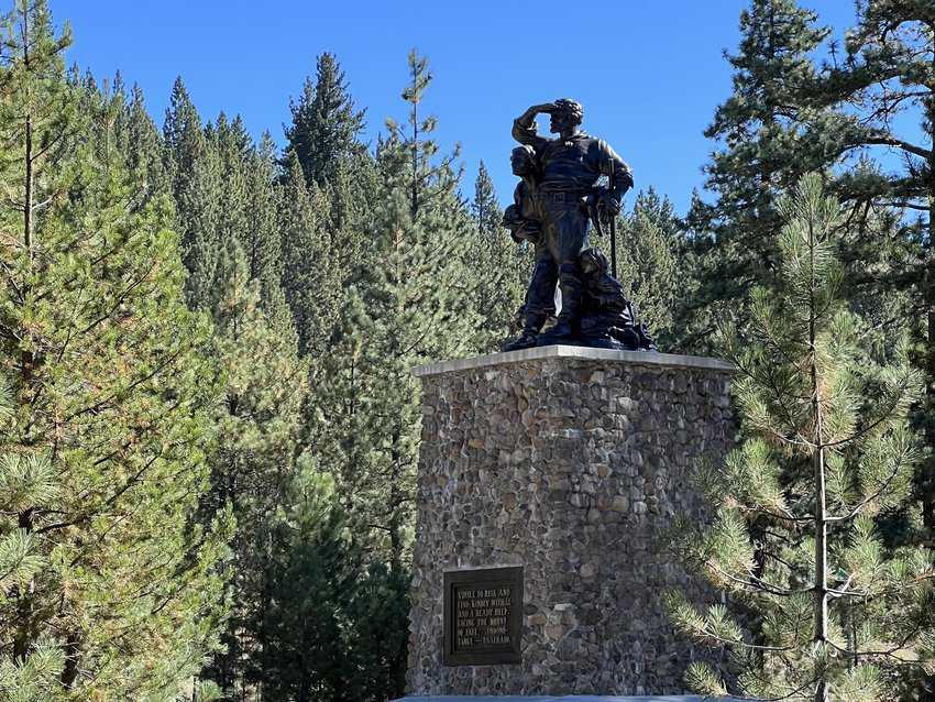 Bronze Image of family looking in the distance atop a stone pedestal surrounded by pine trees and blue sky 