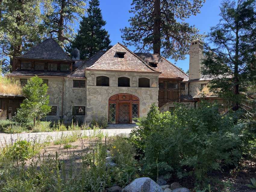 Stone house with large wooden door in front of a drive surrounded by greenery and pine trees. 